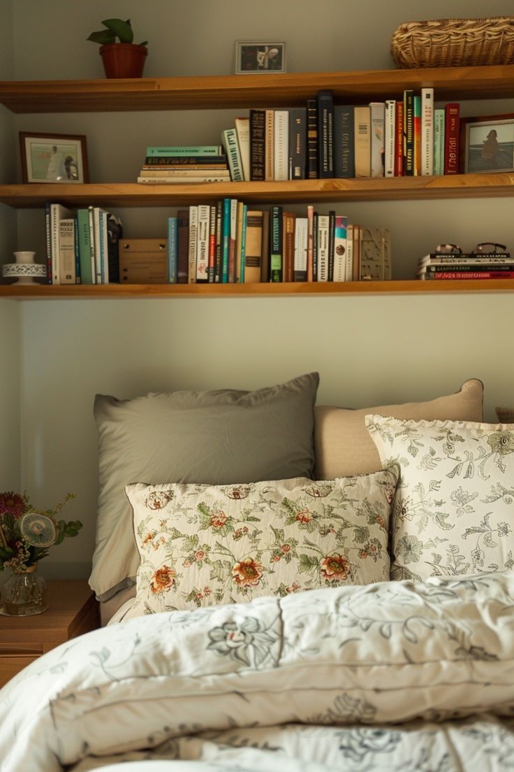 Cozy bedroom with a bookshelf above the bed, decorated with throw pillows and floral bedspread. Space Saving Shelves Bedroom, Wooden Shelves Bedroom Decor, Book Shelf Wall Bedroom, Room Shelving Ideas Bedrooms, Shelf Along Top Of Wall Bedroom, Shelves Books Wall, Drawers With Shelves Above, Wall Long Shelf, High Shelves Bedroom