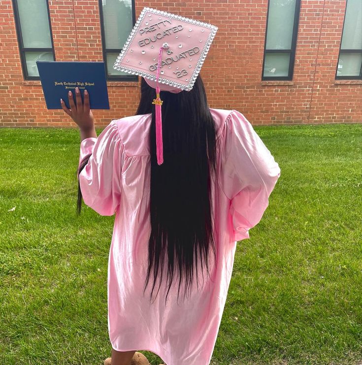 a girl with long black hair wearing a pink graduation gown and holding a blue book