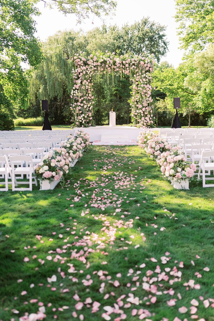 an outdoor ceremony setup with white chairs and pink flowers on the grass in front of it