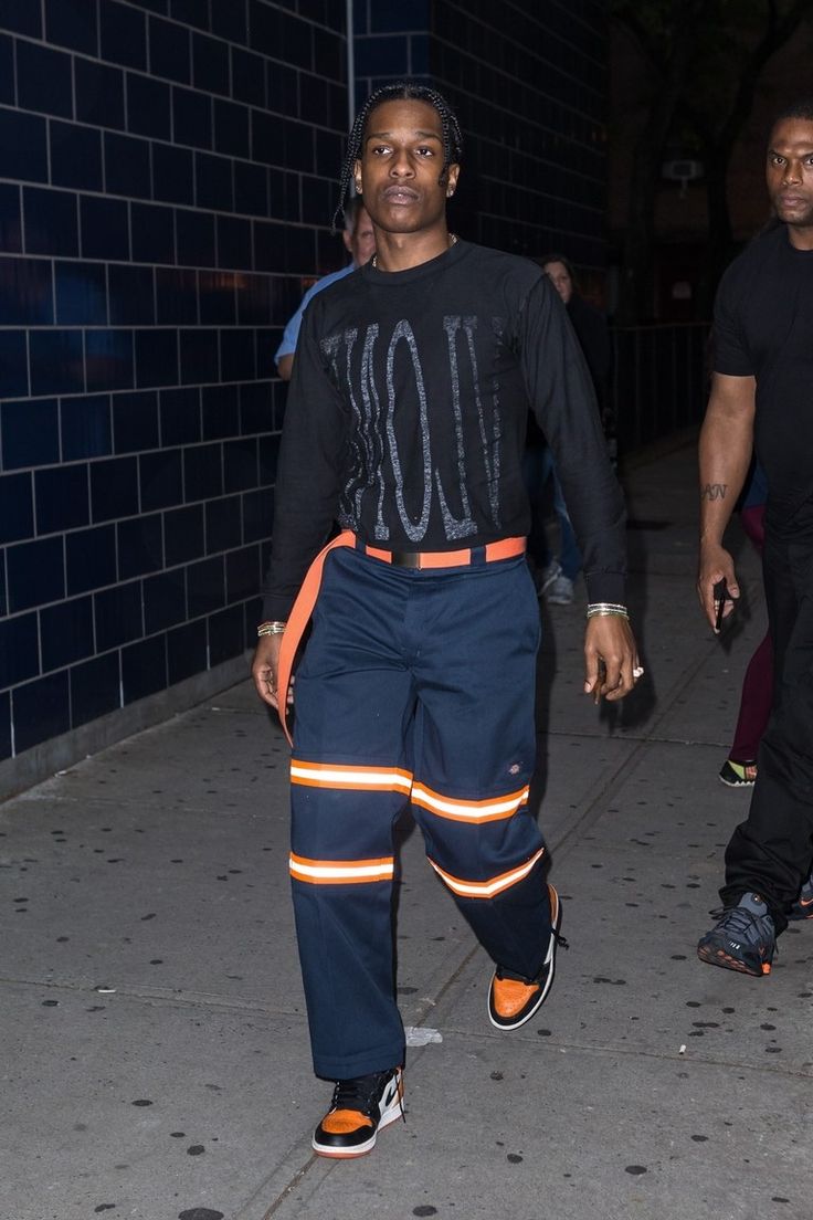 a man in black shirt and blue pants walking on sidewalk next to brick wall at night