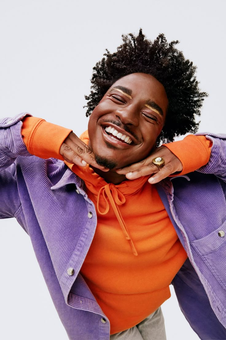 a man in an orange shirt and purple jacket with his hands on his chest smiling