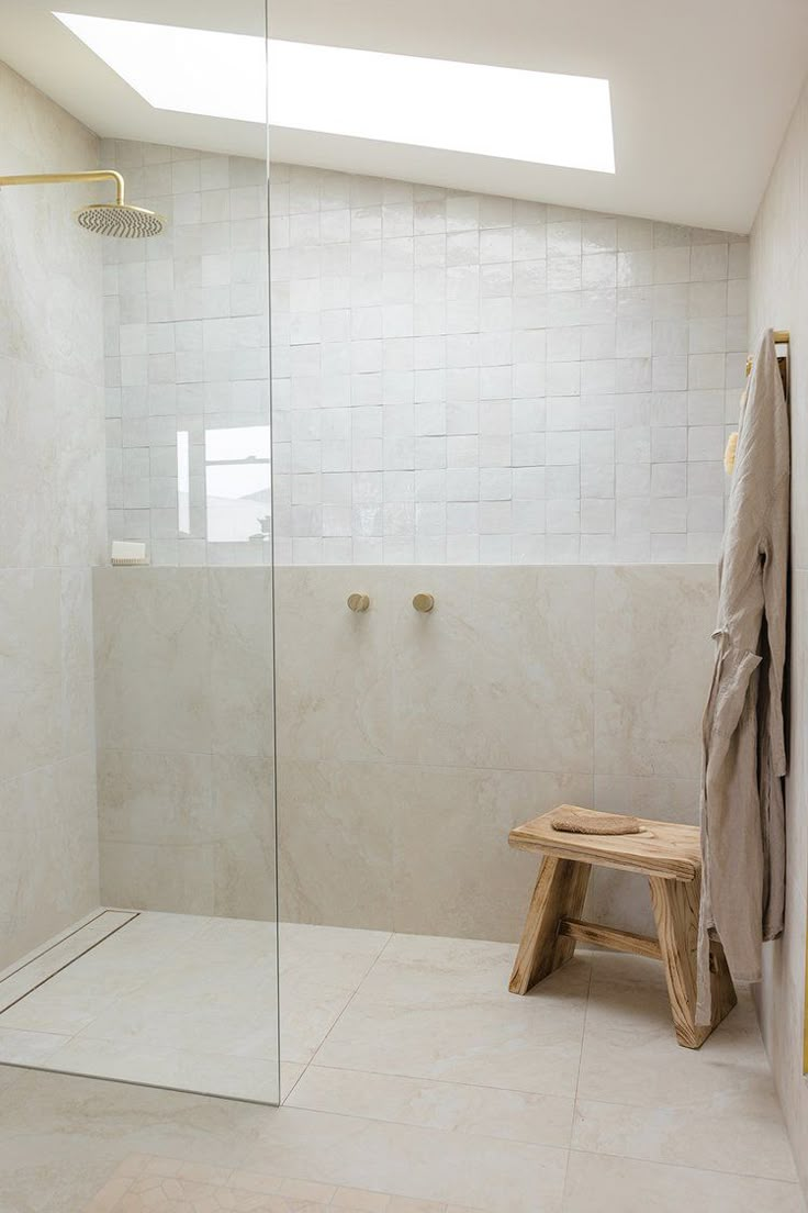 a walk in shower sitting next to a white tiled wall and wooden bench under a skylight