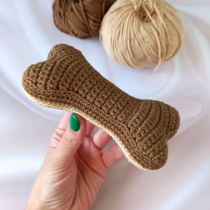 a hand holding a knitted dog bone next to two skeins of yarn