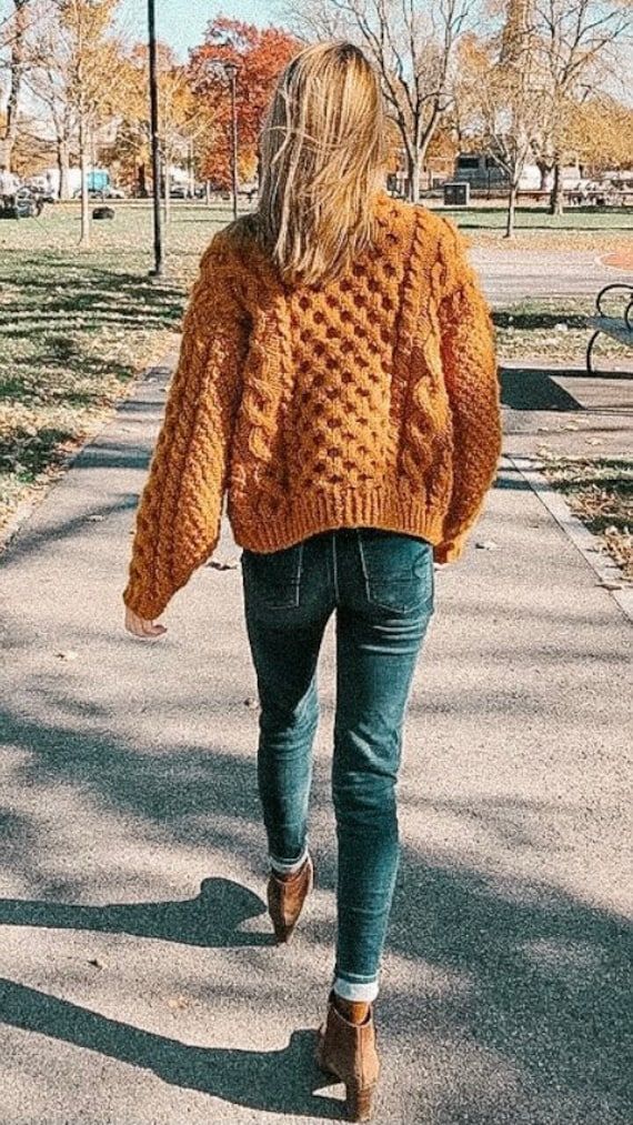 a woman is walking down the sidewalk wearing an orange knitted sweater and blue jeans