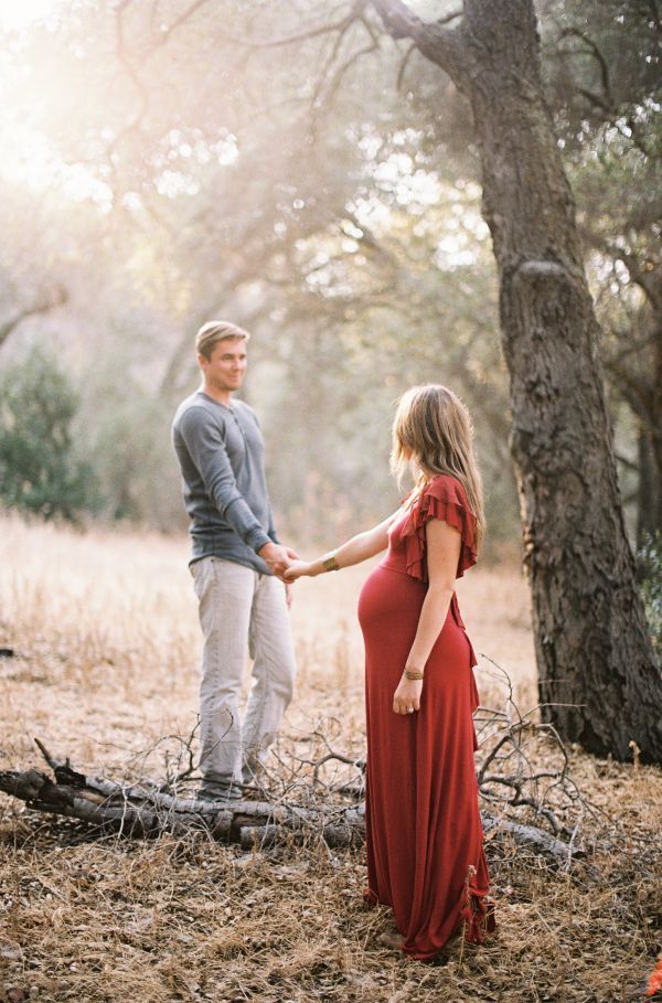 an image of a pregnant couple holding hands and standing in front of a tree with sunlight streaming through the branches