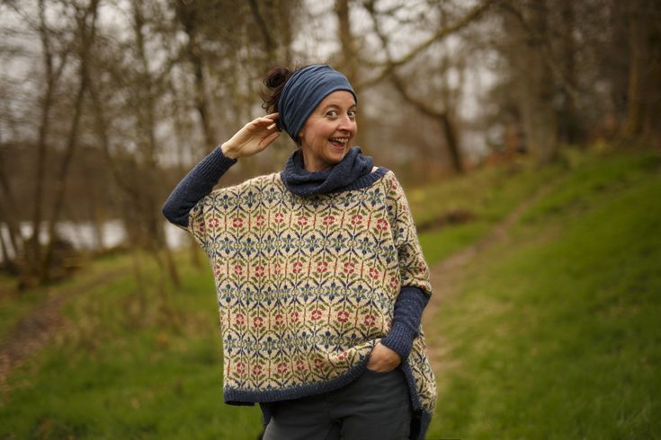 a woman wearing a blue head scarf and a knitted ponchle smiles as she walks down a path