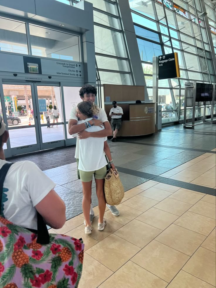 two people hugging each other in front of an airport terminal entrance with glass doors and large windows