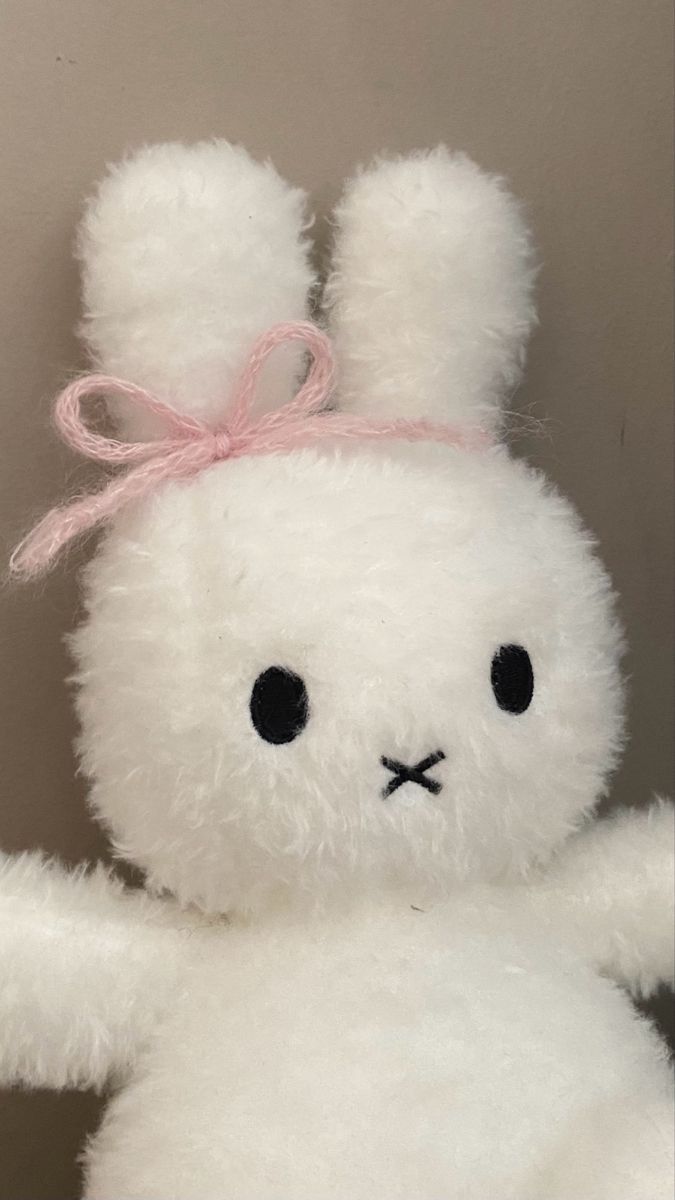 a white stuffed animal with a pink ribbon on its head and ears, sitting in front of a wall