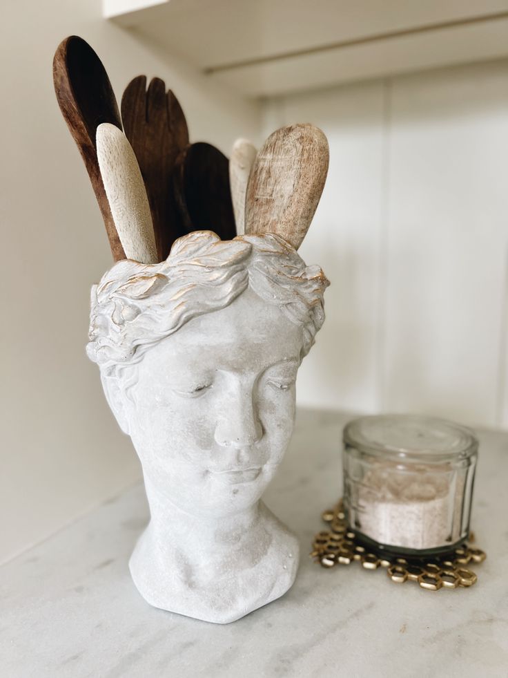 a white statue head sitting on top of a counter next to a jar filled with wooden spoons