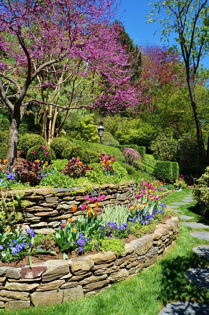 a stone garden wall with flowers and trees in the background, surrounded by green grass