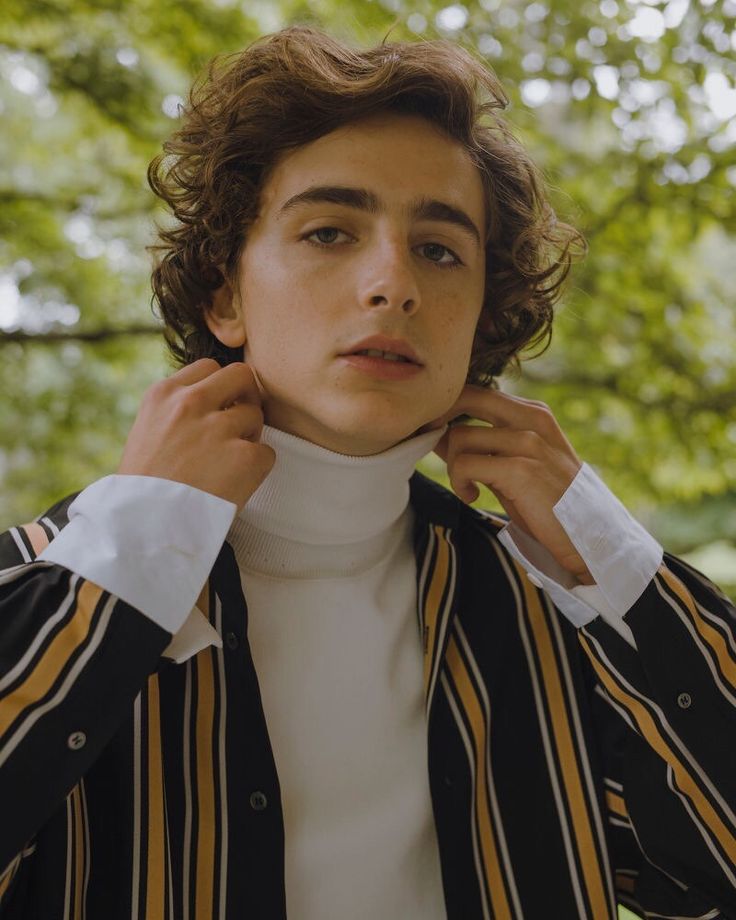 a young man with curly hair wearing a striped jacket and white turtle neck sweater, standing in front of a tree