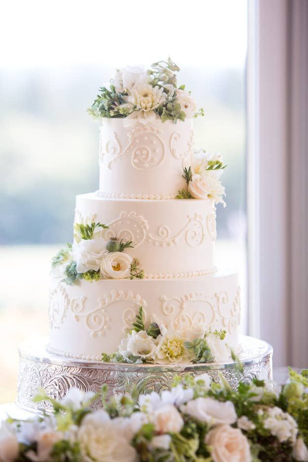 a wedding cake with white flowers and greenery on the top is sitting in front of a window