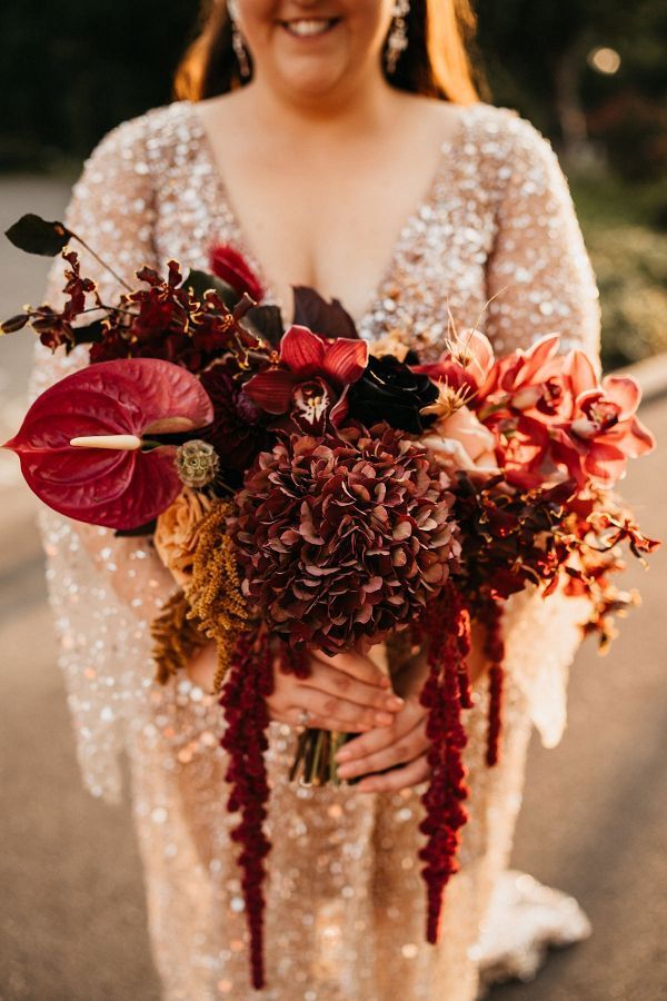 a woman in a white dress holding a red and orange bouquet with greenery on it