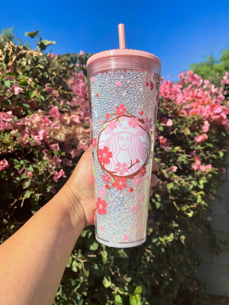 a hand holding a pink starbucks cup in front of some bushes with flowers on it