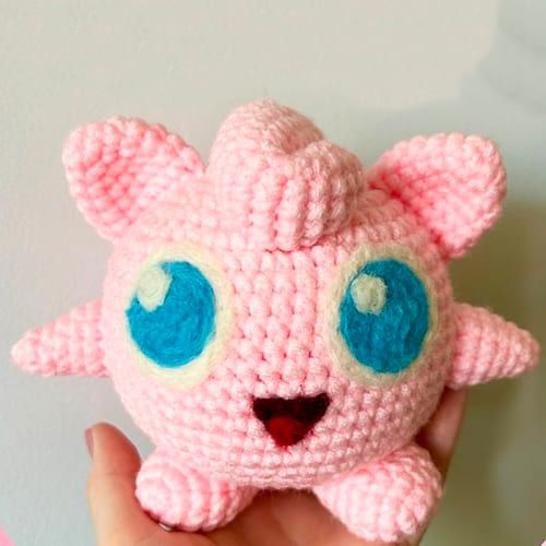 a hand holding a pink crocheted stuffed animal with big blue eyes and ears