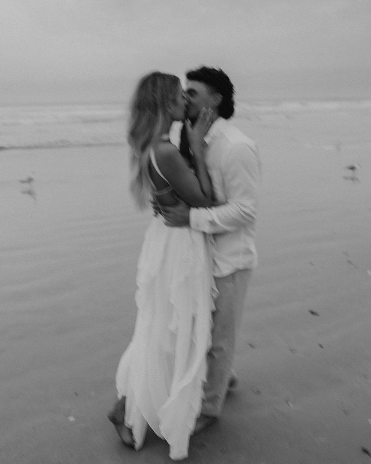 a man and woman kissing on the beach in front of the ocean while holding each other