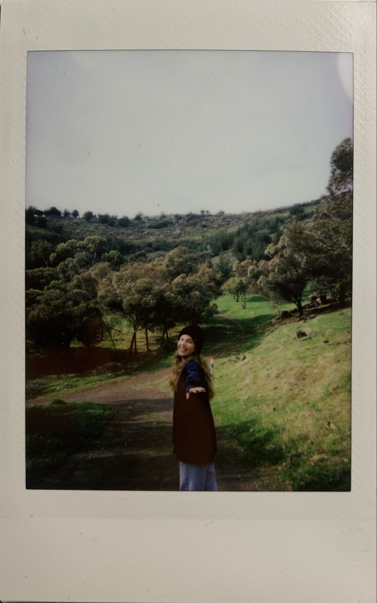 a woman standing in the middle of a field with trees and grass on both sides