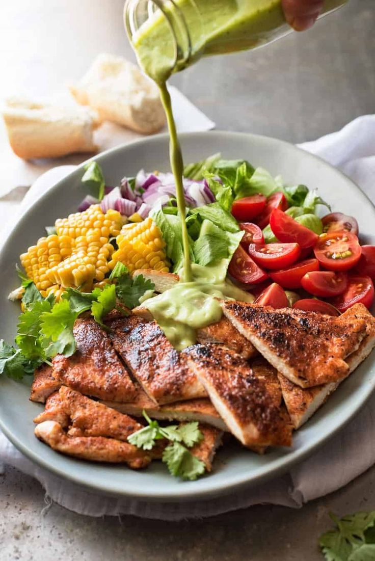 someone pouring dressing onto a plate of chicken, tomatoes, corn and lettuce