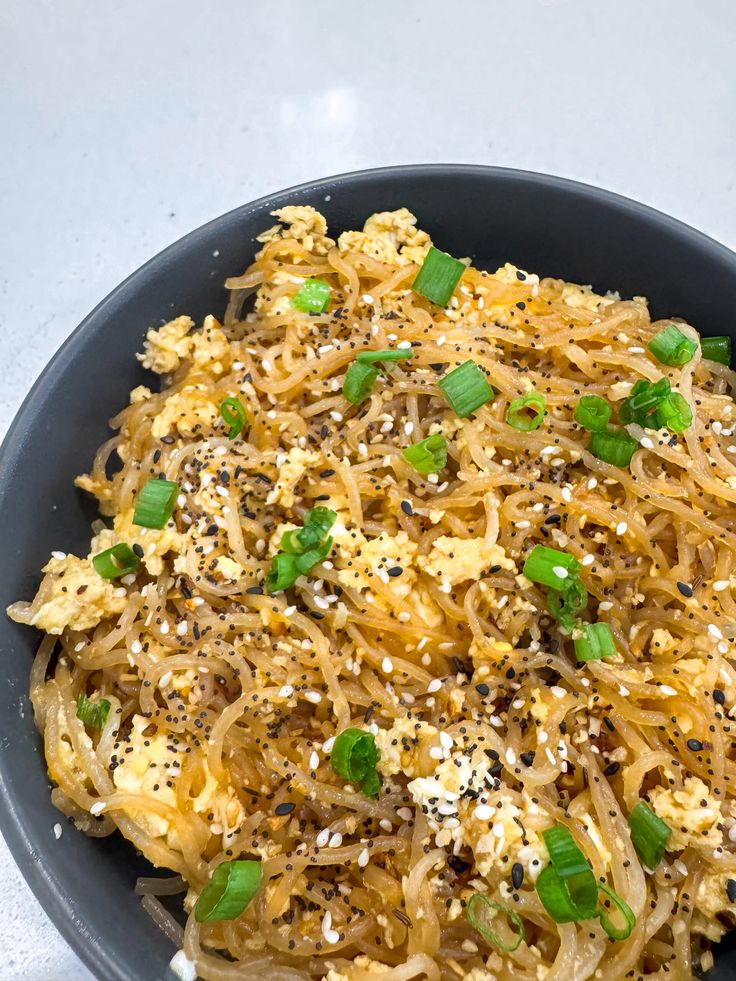 a black bowl filled with noodles and topped with sesame seeds, scallions and green onions