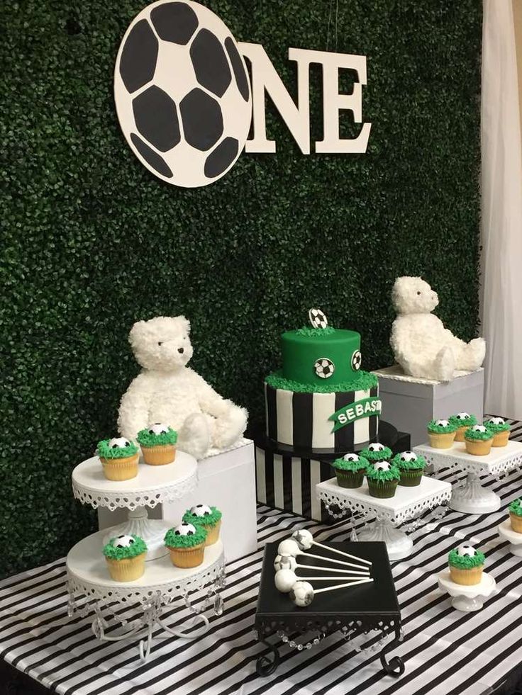 a table topped with cupcakes and teddy bears next to a wall covered in greenery