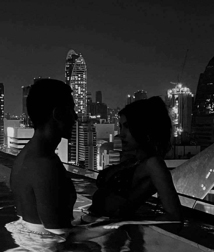 a man and woman are sitting in the pool looking at each other with city lights behind them