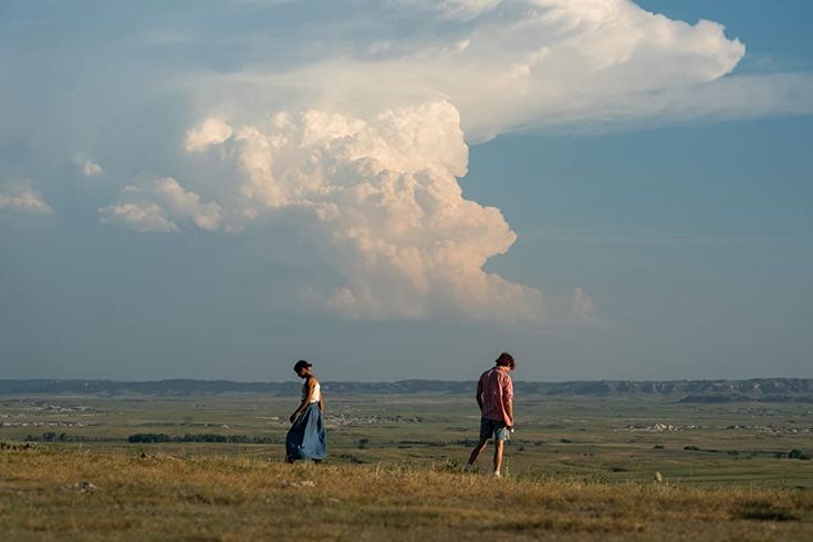 two people standing in a field watching a large cloud loom over the plains behind them