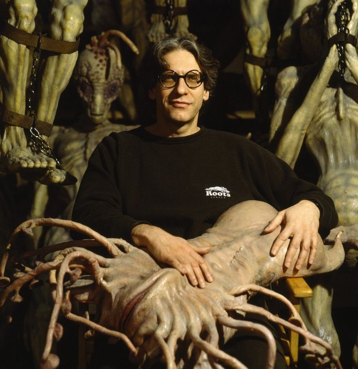 a man with glasses sitting in front of an alien sculpture and looking at the camera