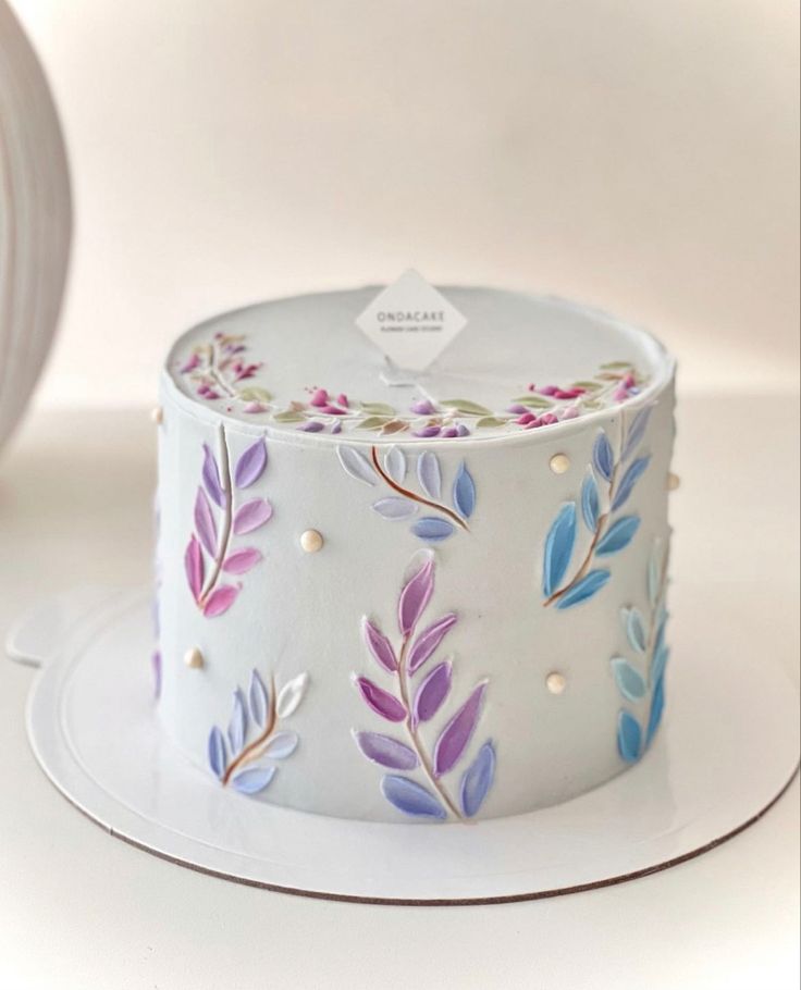 a white cake with purple and blue leaves painted on the top is sitting on a plate