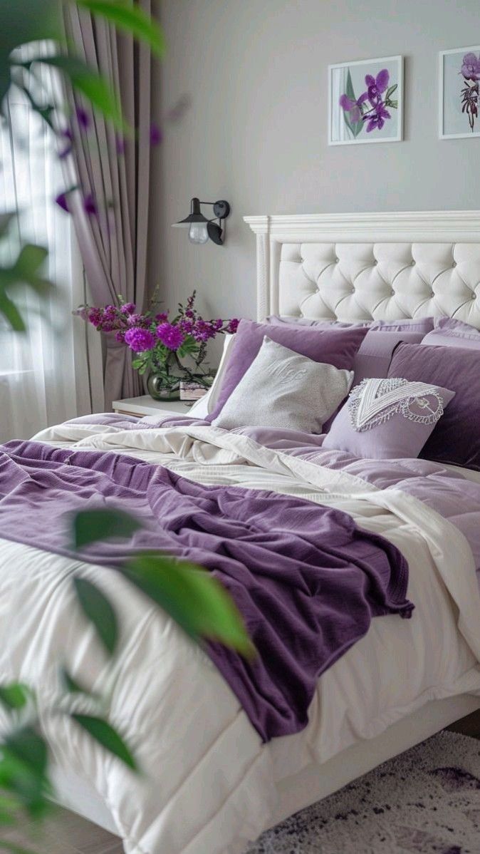 a white bed with purple sheets and pillows in a bedroom next to a potted plant