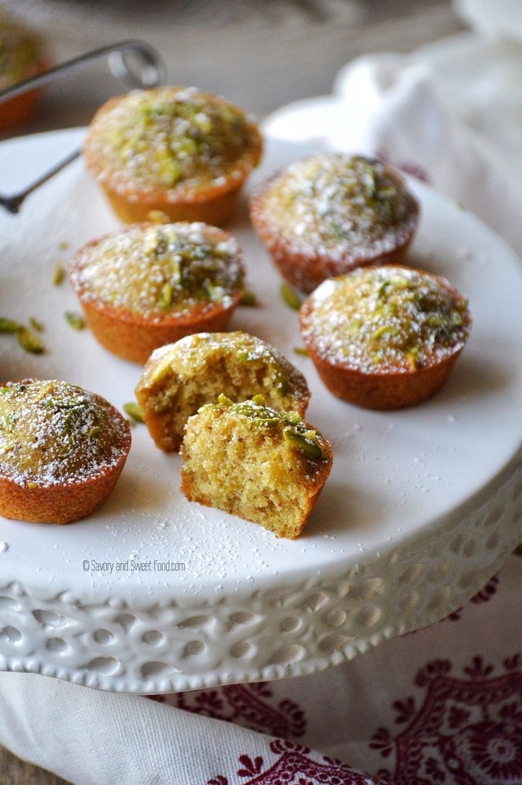 several small muffins on a white plate covered in powdered sugar and sprinkled with pistachio