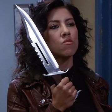 a woman holding a large knife in her right hand and looking at the camera while wearing a brown leather jacket