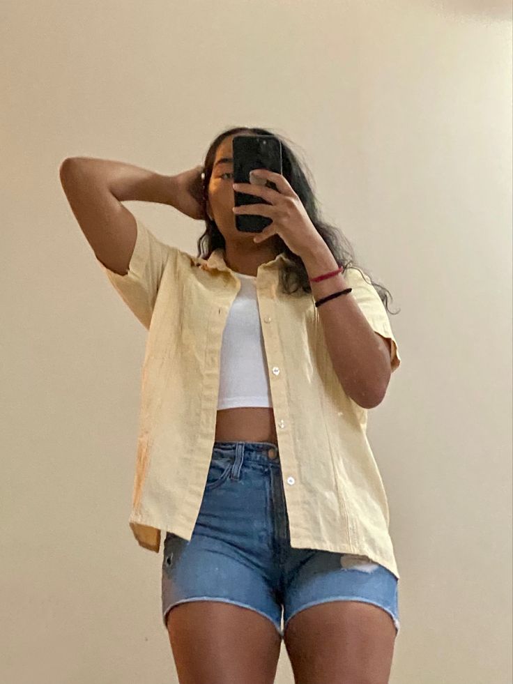 A women taking a mirror selfie of her outfit; a white tank top with a yellow  short sleeve button up and jean shorts. Shorts Top Outfit, Jean Shorts And White Button Up, Polo Shirt And Shorts Women, Button Down And Shorts Women Outfit, Short Button Up Shirt Outfit, Short Sleeve Button Up Shirt Outfit, Short Sleeve Button Up Outfit Women, White Tank Top Outfit Aesthetic, Shorts And Button Up Shirt