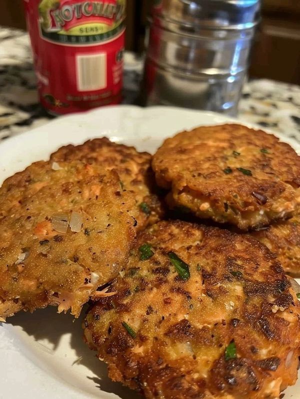 three crab cakes on a white plate next to a can of hot sauce and water