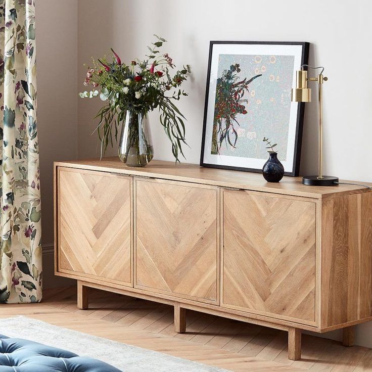 a wooden sideboard with two vases on top and flowers in the corner next to it