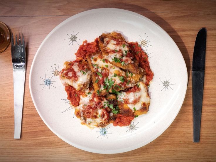 a white plate topped with lasagna next to a fork and knife on top of a wooden table