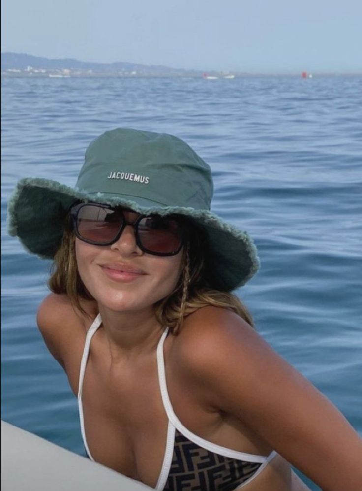 a woman wearing a green hat and sunglasses in the water with boats in the background