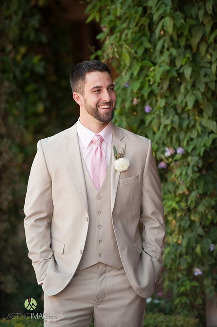 a man in a tan suit and pink tie standing next to a green bush with purple flowers