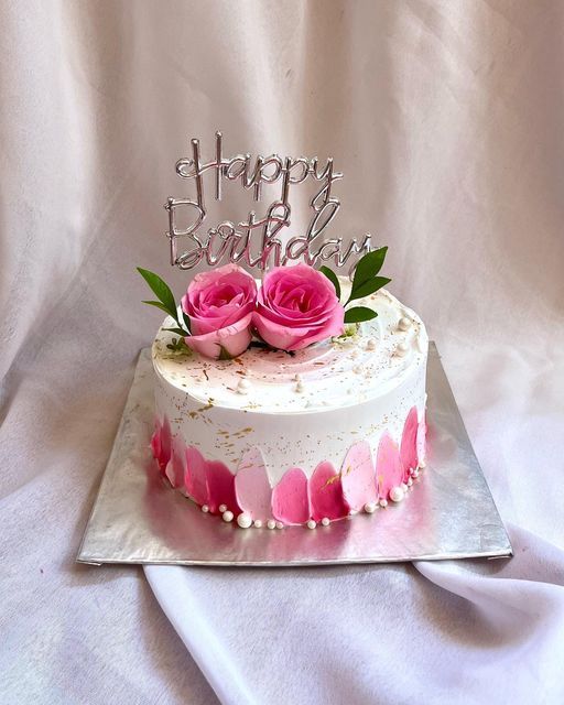 a white cake with pink roses on top and happy birthday written on the side in silver lettering