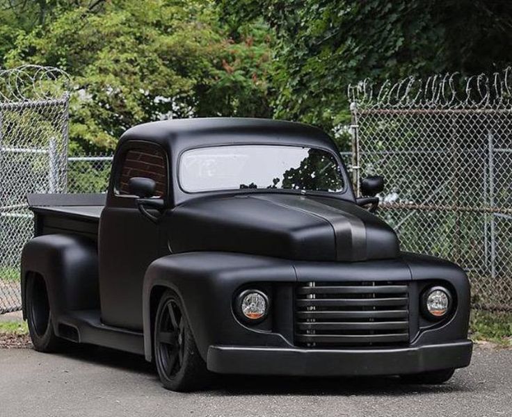 an old black pickup truck parked in front of a chain link fence and tree line