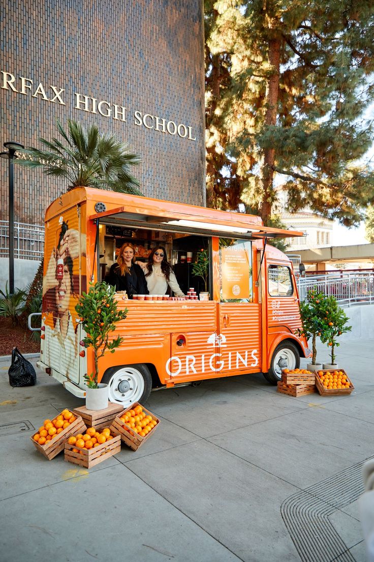 an orange food truck parked in front of a building with people standing outside it and boxes of oranges on the ground