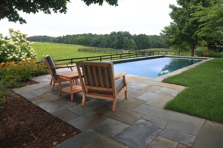 two wooden chairs sitting on top of a stone patio next to a swimming pool in the middle of a lush green field