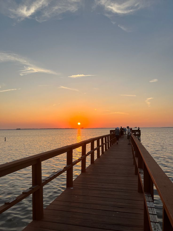 the sun is setting over the water and people are standing on a pier looking out at the ocean