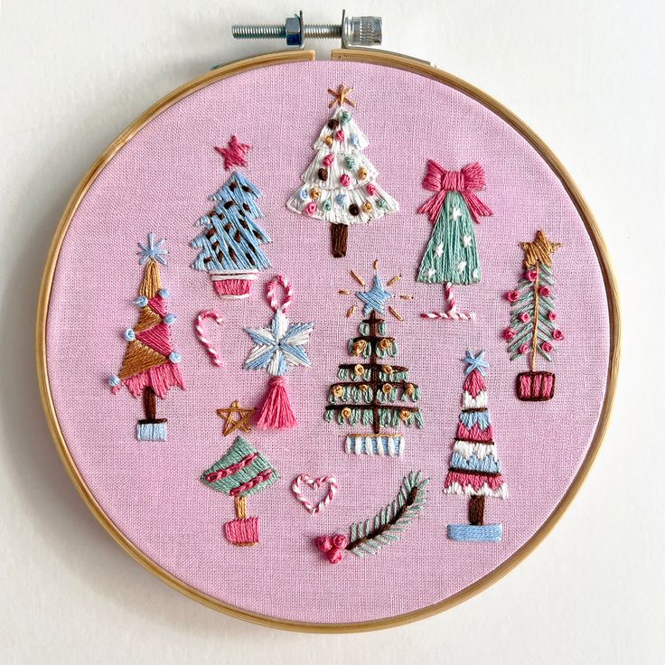 a cross stitch pattern with christmas trees and other decorations on pink fabric in a hoop