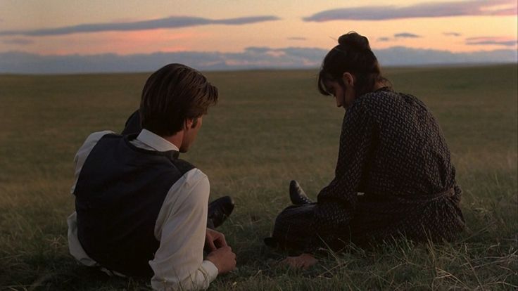 two people sitting on the ground in an open field at sunset, one holding a cell phone