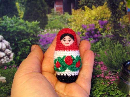 a hand holding a small crocheted doll in front of a garden with flowers