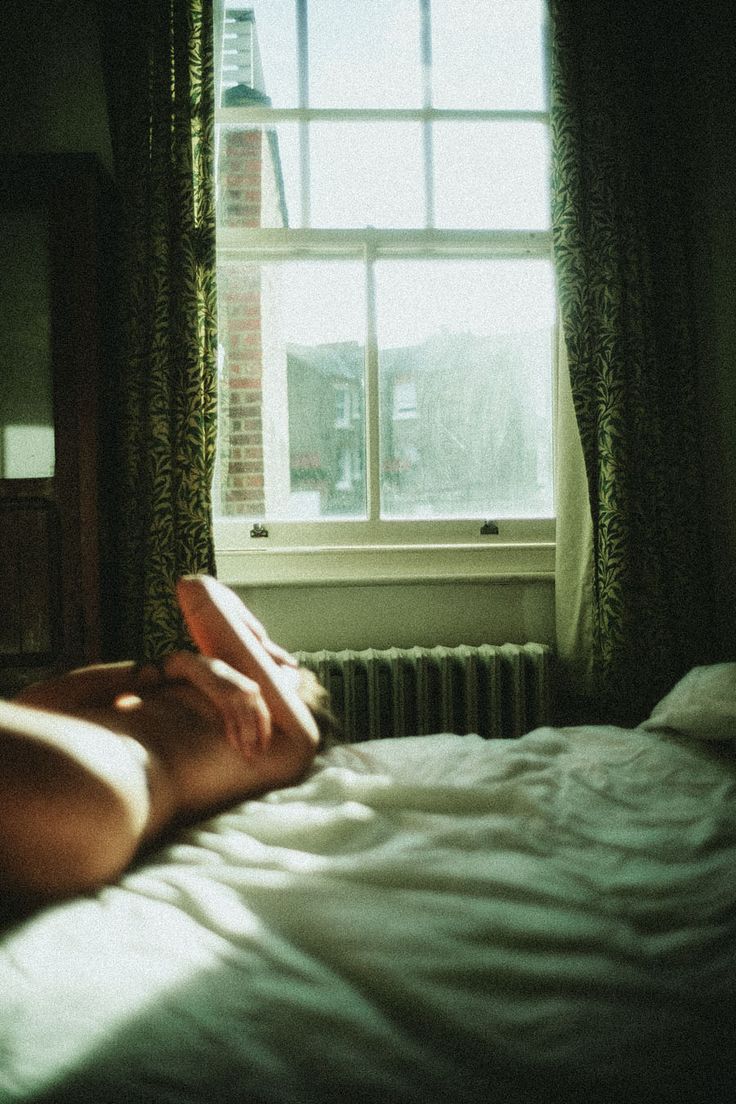 a naked woman laying on top of a bed in front of a window next to a radiator