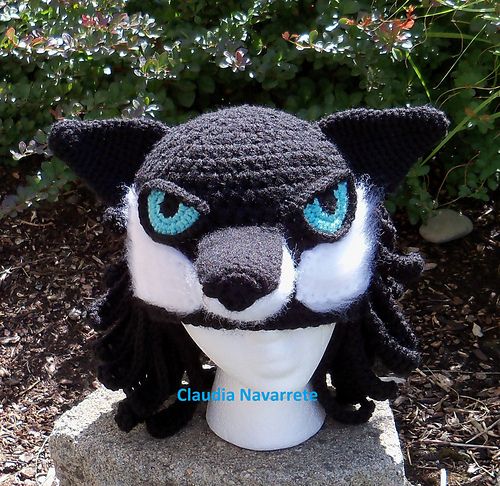 a crocheted black and white wolf head with blue eyes on a rock in front of some bushes