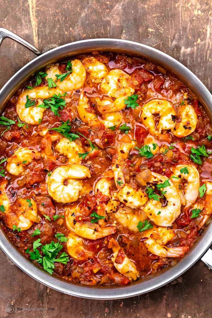 a pan filled with shrimp and tomato sauce on top of a wooden table next to a spoon