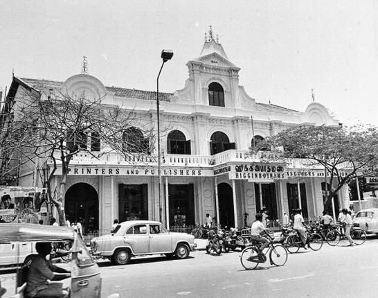 an old black and white photo of people riding bikes in front of a building with cars parked on the street