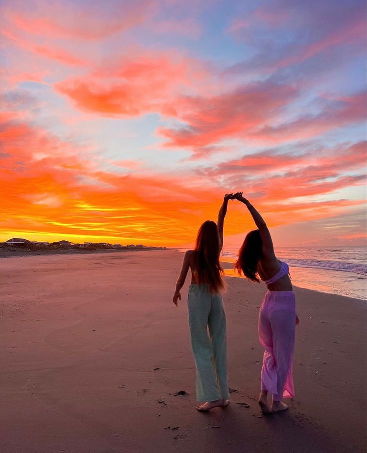 two women standing on top of a beach next to the ocean at sunset with their arms in the air